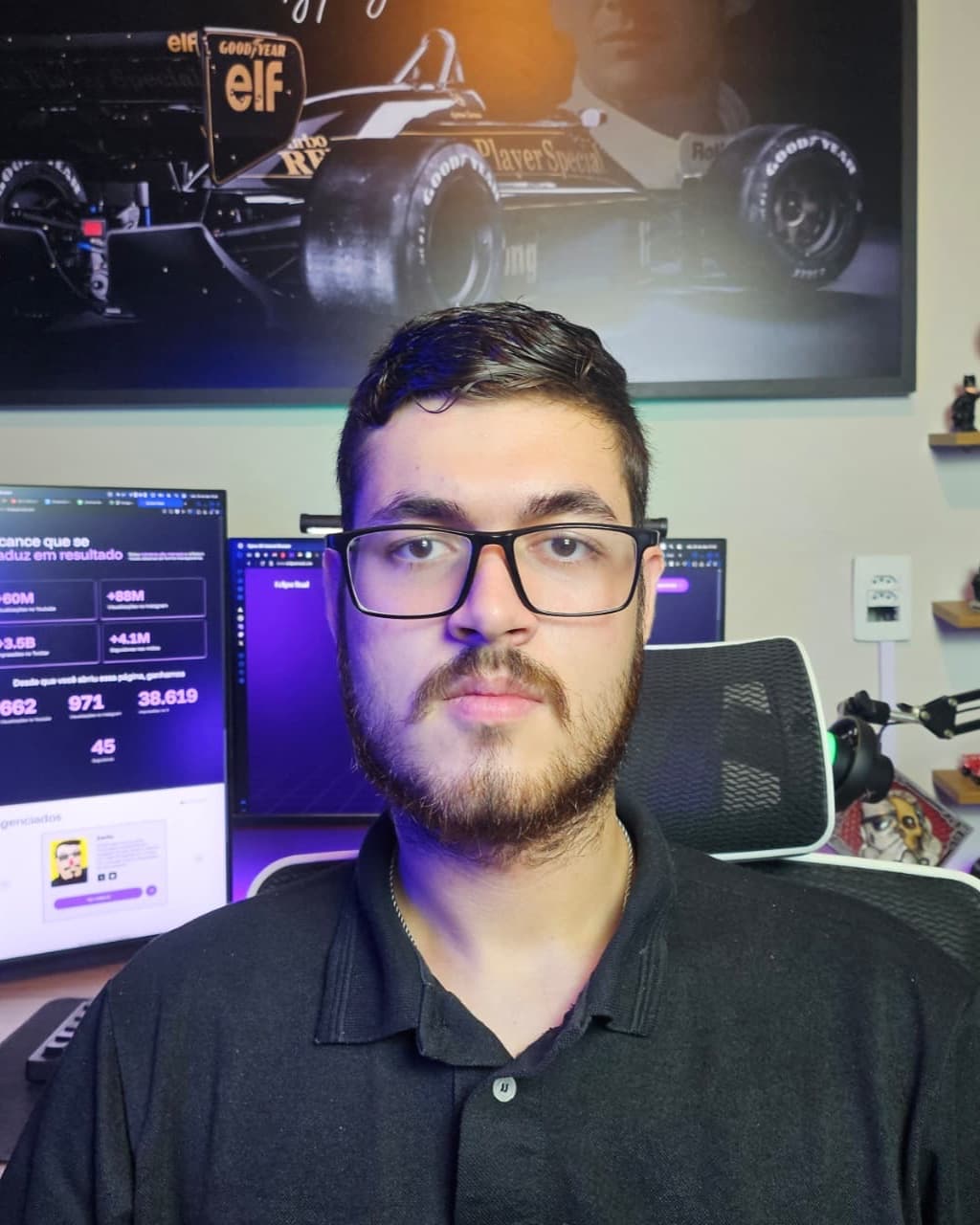 Alex Matida, de óculos, barba e camisa polo preta, em frente aos monitores